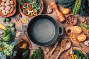 Various organic vegetables ingredients and empty iron cooking pot, wooden bowls, spoons on wooden background. Top view, copy space. Organic vegetables ingredients for vegan cooking. Clean eating food